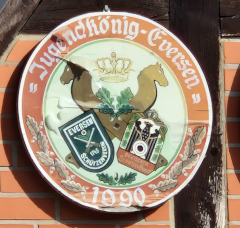 Jugendkönigsscheibe des Schützenvereins Eversen aus dem Jahr 1990. Dargestellt ist zwei zwei stilisierte gekreuzte Pferdeköpfe, das Vereinswappen vom Schützenverein Eversen, eine Krone mittig zwischen den Pferdeköpfen. Das Bild wird umrandet mit der Inschrift Jugendkönig Eversen 1990 Jugendkönigsscheibe des Schützenverein Eversen aus dem Jahr 1990. Dargestellt 