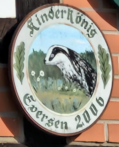 Kinderkönigsscheibe des Schützenvereins Eversen aus dem Jahr 2006: Rundes Schild mit einem gemalten Dachs in einer Wiesenlandschaft, umgeben von Eichenblättern und der Inschrift KinderKönig Eversen 2006.