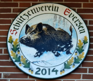 Königsscheibe des Schützenverein Eversen 2014: Runde bemalte Scheibe mit der Aufschrift „Schützenverein Eversen" in gotischer Schrift mit roten Initialen und der Jahreszahl 2014. Im Mittelbild ein handgemalter Keiler im Galopp durch Schnee vor winterlichem Hintergrund. Der Rand ist mit Eichenlaub und Eicheln in Grün und Gelb verziert.