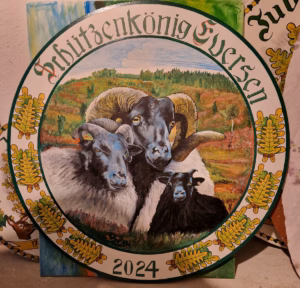Königsscheibe des Schützenkönig Eversen 2024: Runde bemalte Scheibe mit der Aufschrift „Schützenkönig Eversen" und der Jahreszahl 2024. Im Mittelbild eine handgemalte Gruppe schwarzköpfiger Schafe – ein Widder mit markanten geschwungenen Hörnern, ein Mutterschaf und ein Lamm – vor einer herbstlichen Heidelandschaft. Der Rand ist mit Eichenlaub und Eicheln in Grün und Gelb verziert.