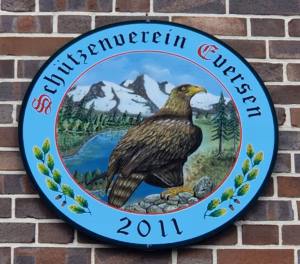 Königsscheibe des Schützenvereins Eversen aus dem Jahr 2011. Rundes Schild mit blauem Hintergrund und handbemaltem Motiv: Ein majestätischer Adler mit gelbem Schnabel und scharfen Krallen steht auf einem Felsen in einer alpinen Landschaft mit schneebedeckten Bergen, einem See und grünen Nadelbäumen. Umrandet von einem Kranz aus grünen Blättern, Eicheln und kleinen Zweigen. Oben der Schriftzug ‚Schützenverein Eversen‘ in gotischer Schrift mit einem roten ‚S‘-Symbol, unten die Jahreszahl ‚2011‘ in schwarzer Schrift.